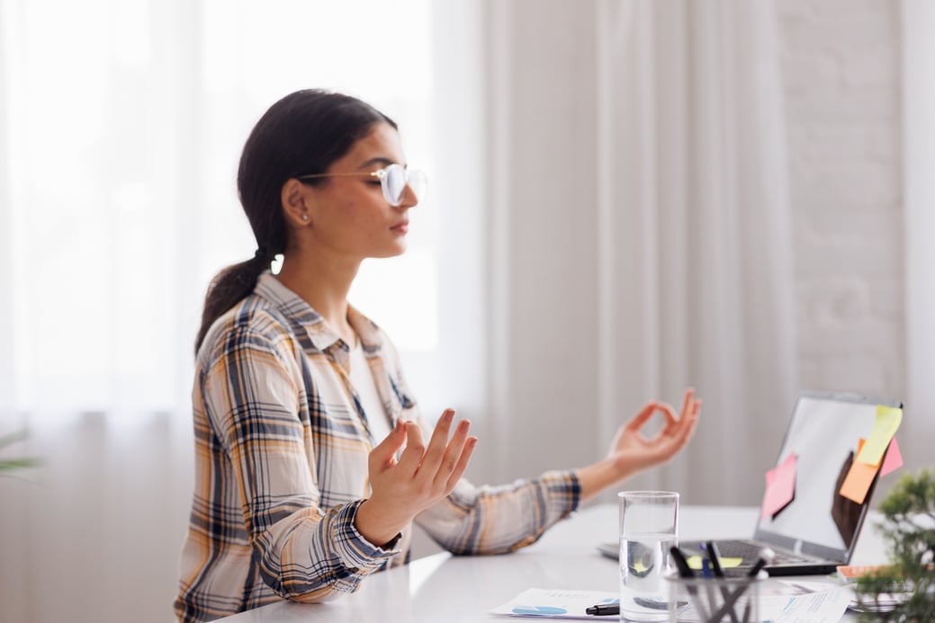 Managing stress in the workplace. Hindu student girl meditating and relaxing during a busy day while sitting at her workplace in an office interior. Focus on the hand.